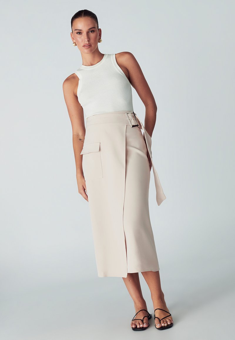 Bela rebrasta brezrokavna majica v kombinaciji s bež visoko pasom zavitim krilom z bočno žepom in podrobnostmi pasu. Črne čevlje na vezanje.