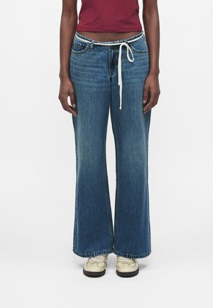 Persoon draagt wijd uitlopende blauwe jeans met een witte koordriem, crèmekleurige sneakers en een cropped bordeauxrode top, staand tegen een effen achtergrond.