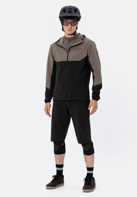 Completo da ciclismo per uomo con giacca leggera nera e grigia, pantaloni corti neri, ginocchiere e casco nero. Tessuto testurizzato e vestibilità atletica.