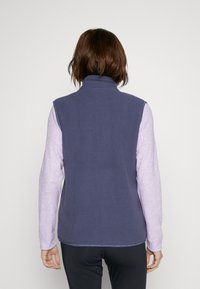 Frau, die eine marineblaue ärmellose Fleece-Weste über einem hellen lila Langarmshirt trägt und mit dem Rücken zu einem schlichten Hintergrund steht.