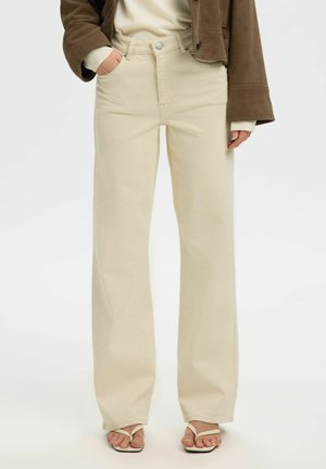 Personne portant un pantalon beige à jambes larges, une veste marron et des sandales blanches à brides, debout, une main dans la poche.