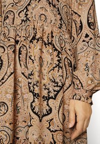 Vestido de manga larga con estampado de paisley en tonos beige, marrón y negro. Presenta puños con volantes y detalle de cintura fruncida.