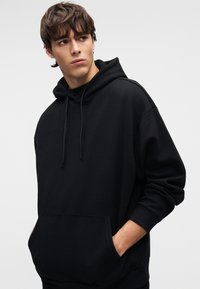 Μαύρη φούτερ με κουκούλα από απαλό ύφασμα, με κορδόνι στην κουκούλα, μεγάλη μπροστινή τσέπη και φαρδιά εφαρμογή με ριμπ μανσέτες. Απλή, χωρίς σχέδια.