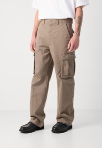 Beige cargo bukser lavet af bomuld, med to sidelommer, to store cargo lommer og et lige ben-design. Båret med sorte sko.