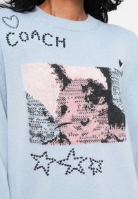 Šviesiai mėlynas megztinis su multicolor katės grafika, juodu užrašu "COACH" ir juoda siuvinėjimo žvaigžde.