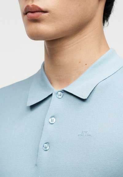 Κοντινό πλάνο ενός ατόμου που φοράει ανοιχτό μπλε πόλο με τρία κουμπιά και ένα μικρό κεντημένο λογότυπο στο στήθος.