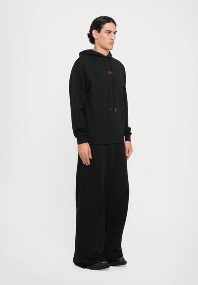 Sweat à capuche noir avec des cordons, et un petit logo. Associé à un pantalon noir ample à jambes larges et des chaussures sombres. Texture lisse.