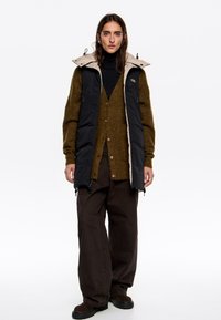 Gilet imbottito nero e beige con colletto alto, indossato sopra un maglione verde oliva, abbinato a pantaloni larghi marrone scuro e stivali neri.