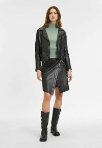 Veste cropped en cuir noir, pull col roulé côtelé vert, jupe enveloppante en cuir noir et bottes noires jusqu'au genou avec des accents en boucle.