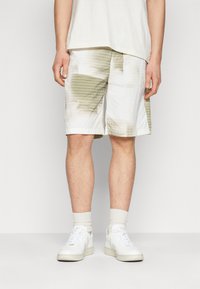 White shorts with green grid patterns and abstract shapes, featuring a relaxed fit and side seam pockets, paired with white sneakers and socks.