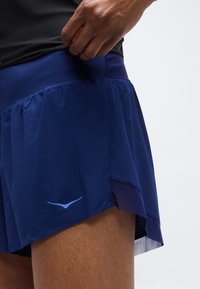 Χέρι που ρυθμίζει τη μέση σκούρων μπλε αθλητικών σορτς με ανοιχτό μπλε λογότυπο στο αριστερό πόδι, μπροστά σε απλό φόντο.