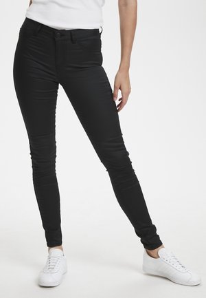 Femme portant un pantalon skinny noir et des baskets blanches, debout, jambes écartées sur un fond blanc uni.