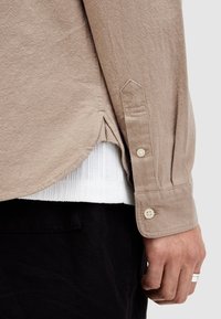 Camisa texturizada beige de manga larga, con dobladillo redondeado, aberturas laterales y cierres de botón en los puños, superpuesta sobre una camiseta blanca de canalé.