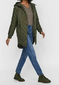 Prešívaná olivově zelená bunda s kapucí a dlouhými rukávy, se dvěma kapsami na zip, v kombinaci se světle modrými džíny a tmavě zelenými šněrovacími teniskami.