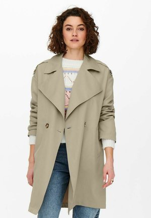 Beige trenchcoat med brede klapper, dobbeltradede knapper og skulderstropper, parret med en lys sweater og blå jeans.