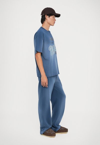 Mladý muž v modrom nadmernom tričku a nohaviciach, s tmavohnedou čiapkou a hnedými vzorovanými slip-on topánkami, stojaci v profilu.