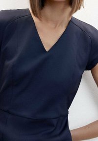 Robe bleu marine avec un décolleté en V et des manches courtes, présentant un tissu lisse et des détails de couture subtils. Sa coupe ajustée met en valeur la silhouette.