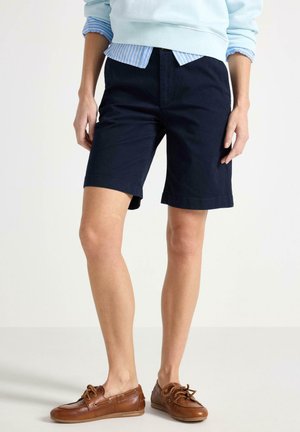 Person iført mørkeblå knælange shorts, lyseblå sweater med stribet skjorte under, og brune læder bådsko.