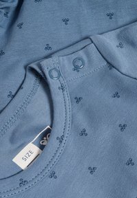 Blå bomullsskjorta med rund halsringning, prydd med små mörkblå blommönster, tryckknappar och ett tygmärke för storleksinformation.