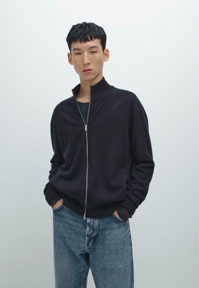 Junger Mann mit kurzen schwarzen Haaren trägt einen dunklen Reißverschluss-Pullover über einem schwarzen Shirt und blauen Jeans vor einem schlichten hellen Hintergrund.