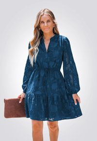 Abito blu navy con texture floreale, scollatura a V, maniche lunghe a sbuffo e vita cintrata. La modella tiene in mano una pochette marrone.