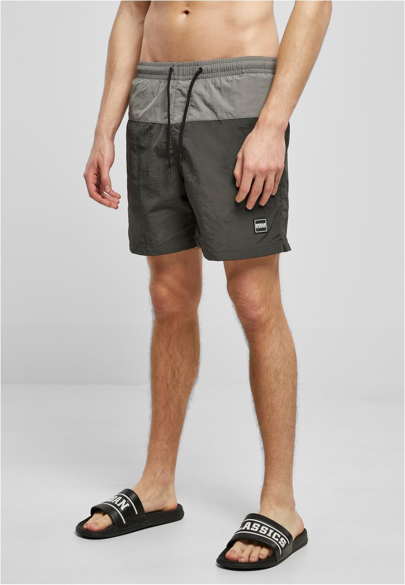 Graue Badehose mit elastischem Bund, Kordelzug und einem Logopatch. Kombiniert mit schwarzen Slide-Sandalen mit weißem, bedrucktem Riemen.