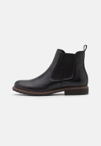 Bottine Chelsea en cuir noir avec panneau latéral élastique, couture de welt marron, tirette au talon et semelle empilée basse sur fond blanc.