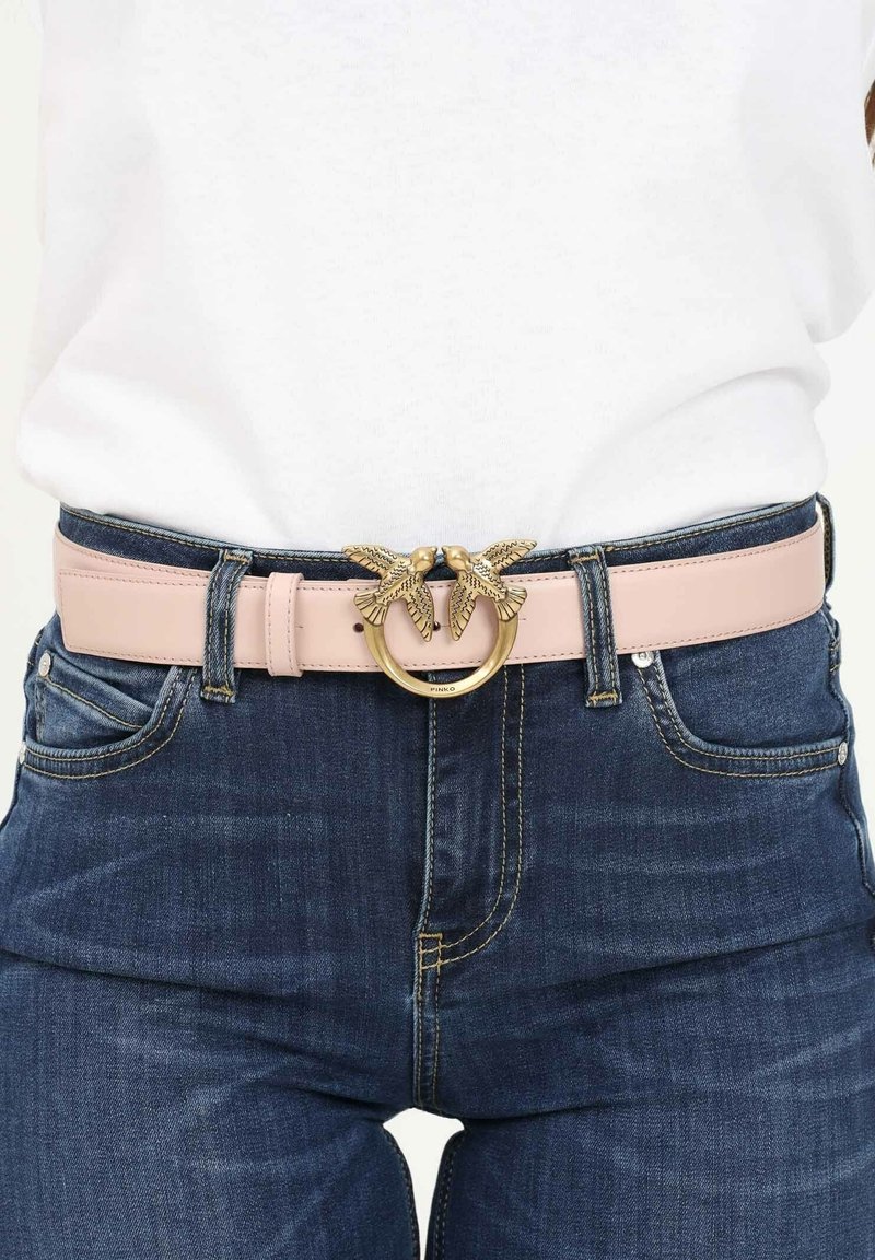 Ceinture en cuir rose clair avec bouclerie dorée en forme de deux oiseaux sur un jean bleu foncé taille haute et un haut blanc.