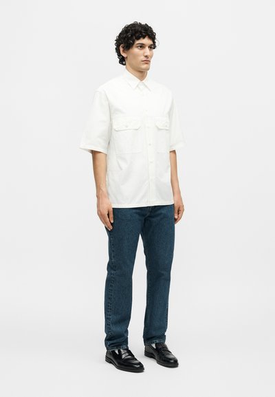 Άνδρας με σγουρά μαλλιά που φοράει λευκό πουκάμισο με κοντά μανίκια και κουμπιά, μπλε τζιν και μαύρα δερμάτινα παπούτσια, στέκεται μπροστά σε ένα μονόχρωμο φόντο.