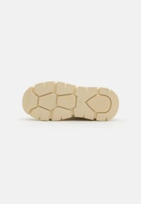 Beige rubber schoenzool met een chunky profiel; beschikt over hexagonale inkepingen voor grip en een gladde, afgeronde teen.