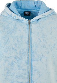 Lys blå zip-up hettegenser i et falmet, marmorert mønster. Har en frontglidelås og en stor hette. Laget av myk, teksturert stoff.