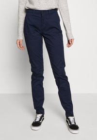 Námornícko modré slim-fit nohavice vyrobené z bavlny, s klasickým zapínaním na gombík, obsahujúce bočné vrecká a hladkú štruktúru. Skombinované s čiernymi teniskami.