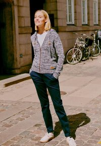 Strikket grå zip-up hettegenser med to treknapper, kombinert med marineblå bukser og hvite sneakers, plassert mot en brosteinsgate.