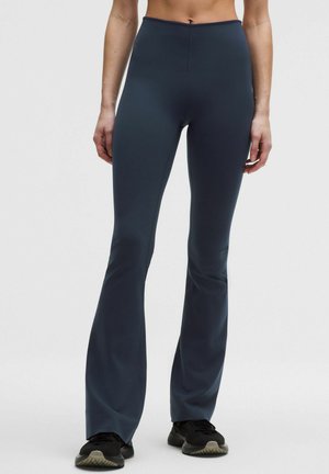 Žena v tmavomodrých rozšírených nohaviciach s vysokým pásom a čiernych teniskách stojí na jednoduchom svetlom pozadí.