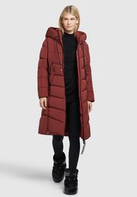 Cappotto lungo imbottito color burgundy con cappuccio, dotato di chiusura con zip, due tasche laterali e stringhe nere. Indossato sopra un top scuro e leggings.