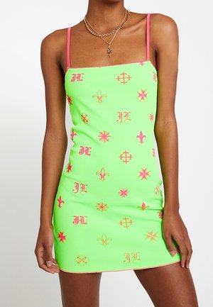 Grünes figurbetontes Minikleid mit dünnen rosa Trägern und roten sowie rosa Blumen- und Emblem-Mustern, kombiniert mit geschichteten goldenen Halsketten.