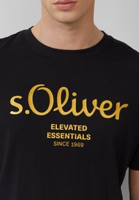 Schwarzes Baumwoll-T-Shirt mit goldenem Text: "s.Oliver" in Schreibschrift, "ELEVATED ESSENTIALS" und "SINCE 1969" in fetter Schrift. Rundhalsausschnitt, kurze Ärmel.