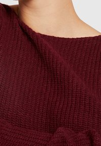 Burgunderfarbener Strickoberteil mit gerippter Textur und weitem Ausschnitt. Der Stoff wirkt weich und gemütlich und lädt zu einer Nahaufnahme ein.