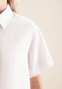 Camicia bianca in lino a maniche corte con colletto e superficie testurizzata. Presenta linee pulite e una vestibilità rilassata.