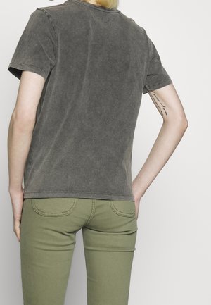 Osoba z krótkimi blond włosami, ubrana w wyblakły czarny t-shirt i oliwkowe spodnie, pokazująca tatuaż na lewym górnym ramieniu.