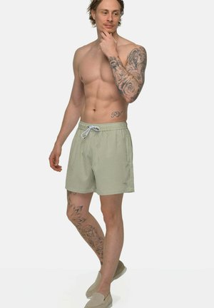 Man zonder shirt met tatoeages op arm en been, die lichtgroene gestreepte shorts en beige instapschoenen draagt, staand met hand op kin.