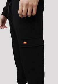 Pantaloni cargo neri con un design aderente, dotati di una tasca laterale decorata con un logo colorato. Tessuto testurizzato con dettagli di cucitura visibili.