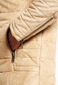 Giacca imbottita beige con una texture morbida, dotata di una tasca laterale con zip e un motivo trapuntato su corpo e maniche.