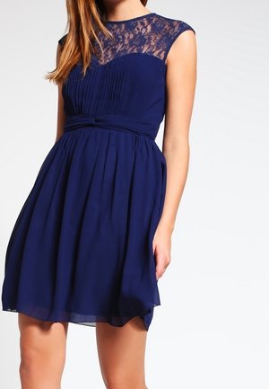 Robe bleu marine sans manches avec corsage plissé, empiècement en dentelle, taille ajustée et jupe fluide, portée par une femme à la peau claire et aux cheveux bruns.