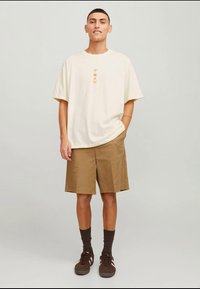 Világos bézs színű, túlméretes póló kis narancssárga grafikával; khaki rövidnadrág; barna sportcipő; sötét zokni. Egyszerű, kényelmes, lezser öltözék, laza szabás.