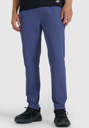 Mężczyzna ubrany w niebieskie spodnie sportowe i czarne buty sportowe stoi na tle jednolitego tła.