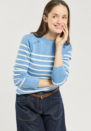 Jeune femme souriante, main sur la joue, portant un pull rayé bleu et blanc et un jean foncé avec une ceinture marron tressée.