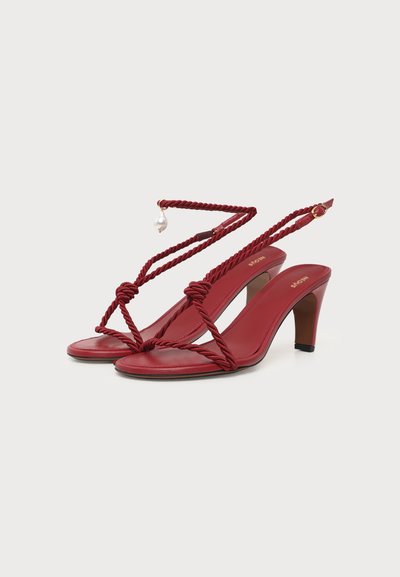 Sandales à talon rouges avec brides en corde torsadée, boucle à la cheville et pendentif perle en détail, présentées sur fond blanc uni.