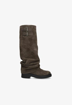 Botte haute en daim marron avec tige souple, deux sangles à boucle métallique réglables près du haut, et une semelle noire à petit talon.