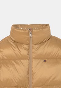 Tan pufferjakke med høj krave, fuld lynlåslukning og logodetalje. Materialet er glat og skinnende med quiltet tekstur.